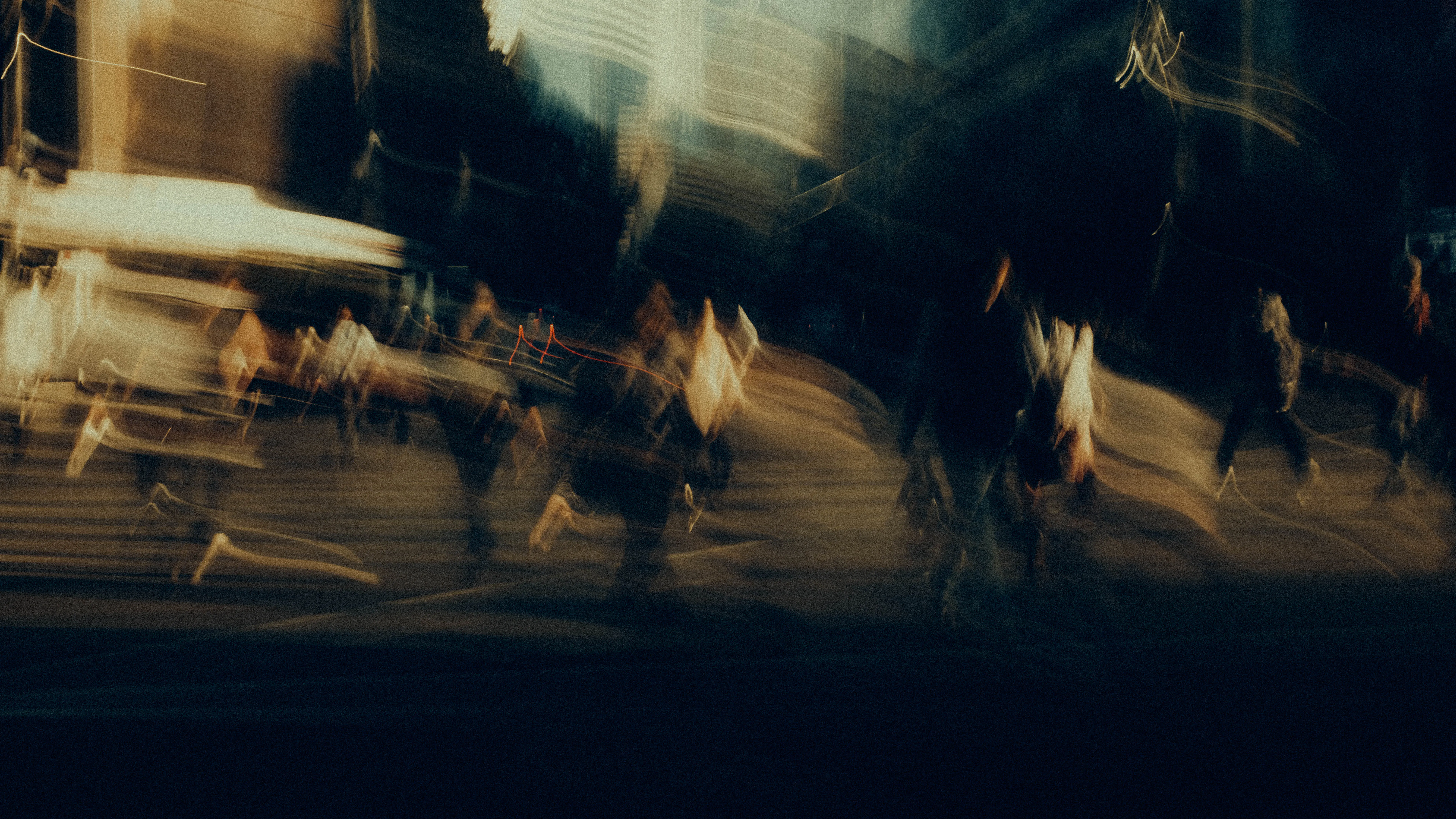 A crowd streams across a night intersection in warm amber motion blur, their figures dissolving into layered streaks of light and shadow against the deep dark of the surrounding city.