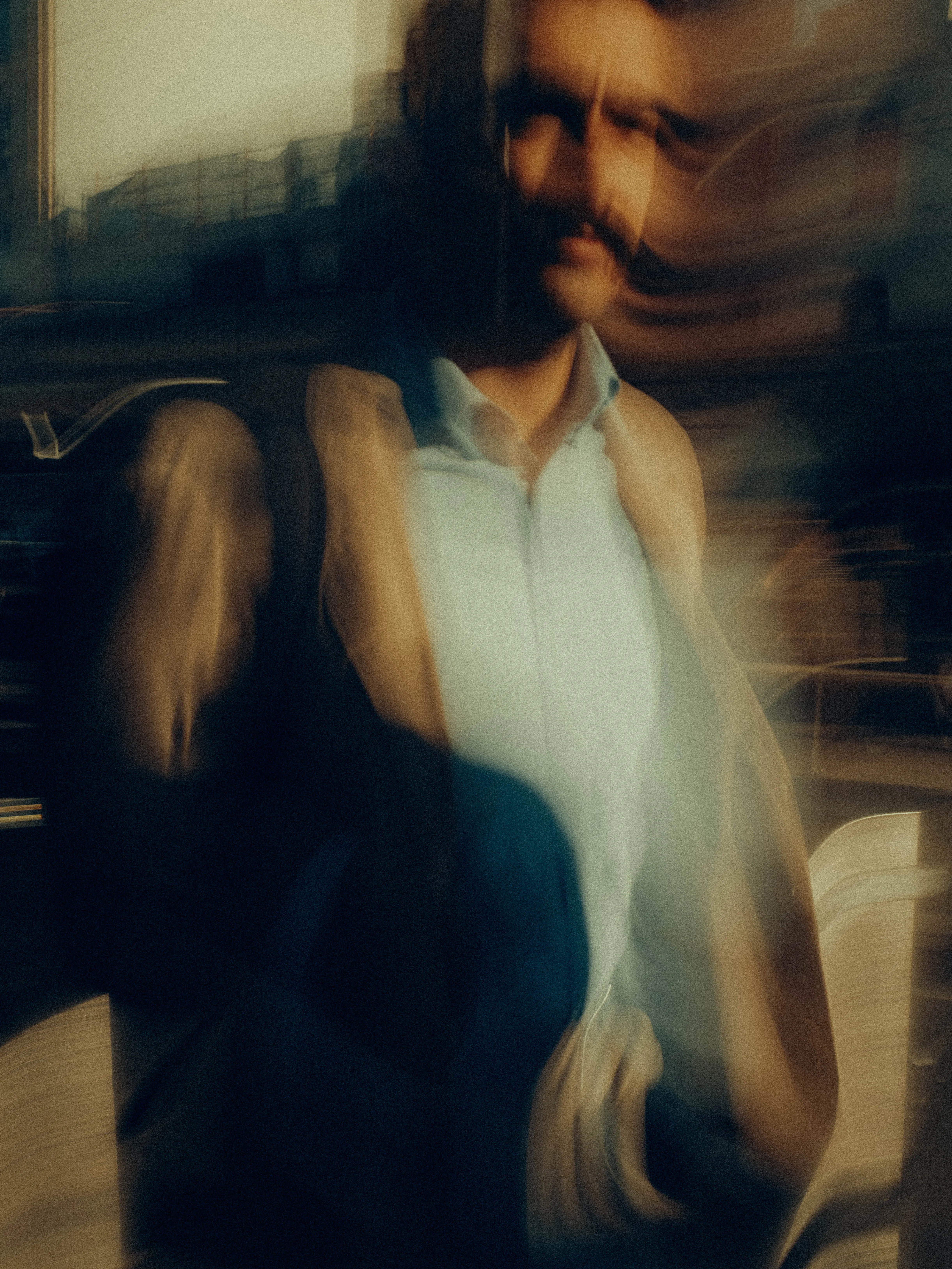 A bearded man in a pale open shirt and dark jacket blurs into warm amber dusk, his silhouette multiplied and fractured against a distant city skyline in rich cinematic tones.