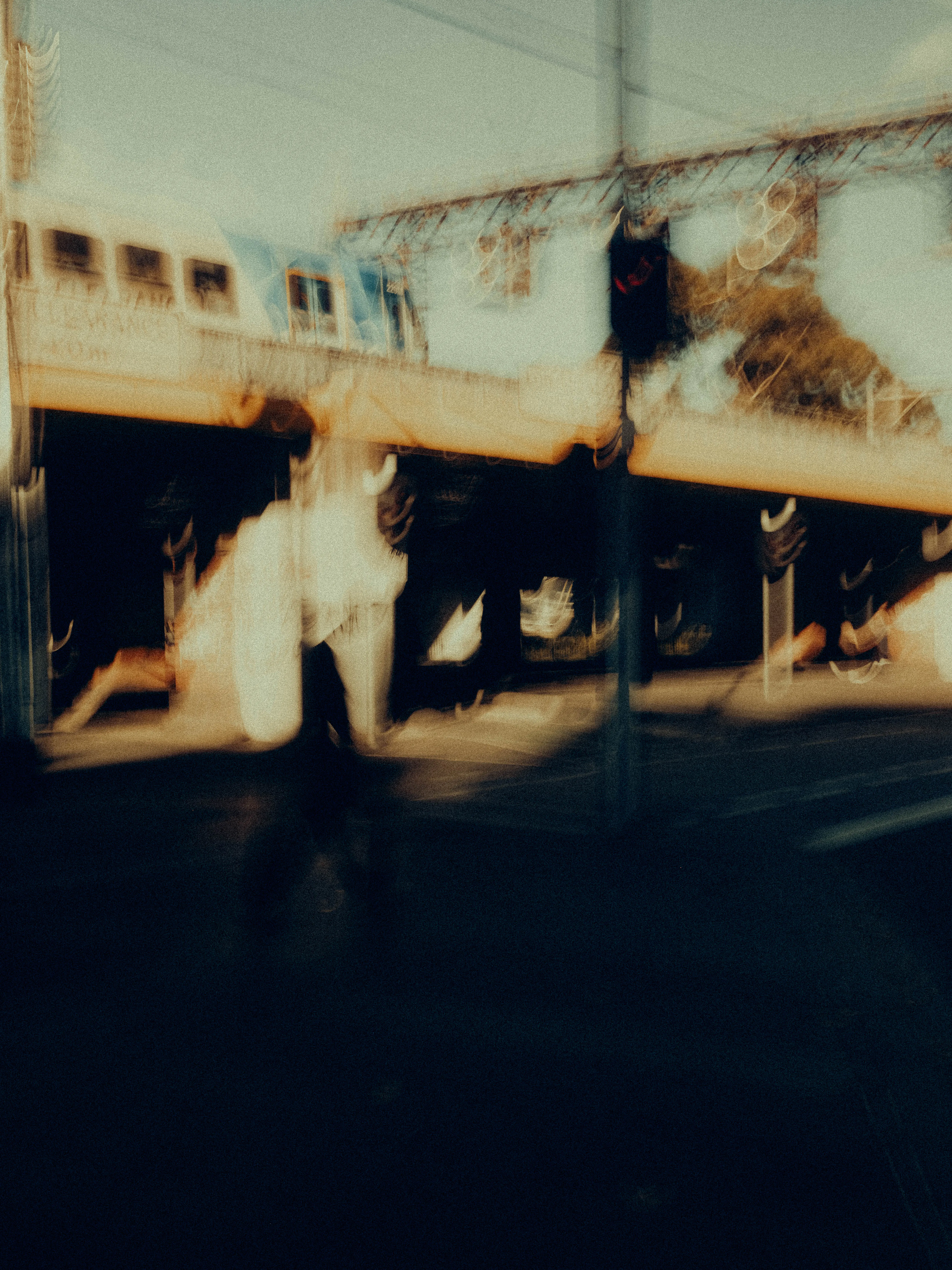 A lone figure in white strides beneath a Melbourne train overpass as a blue train crosses overhead, the scene washed in golden late-afternoon light with long shadows pooling across the road below.