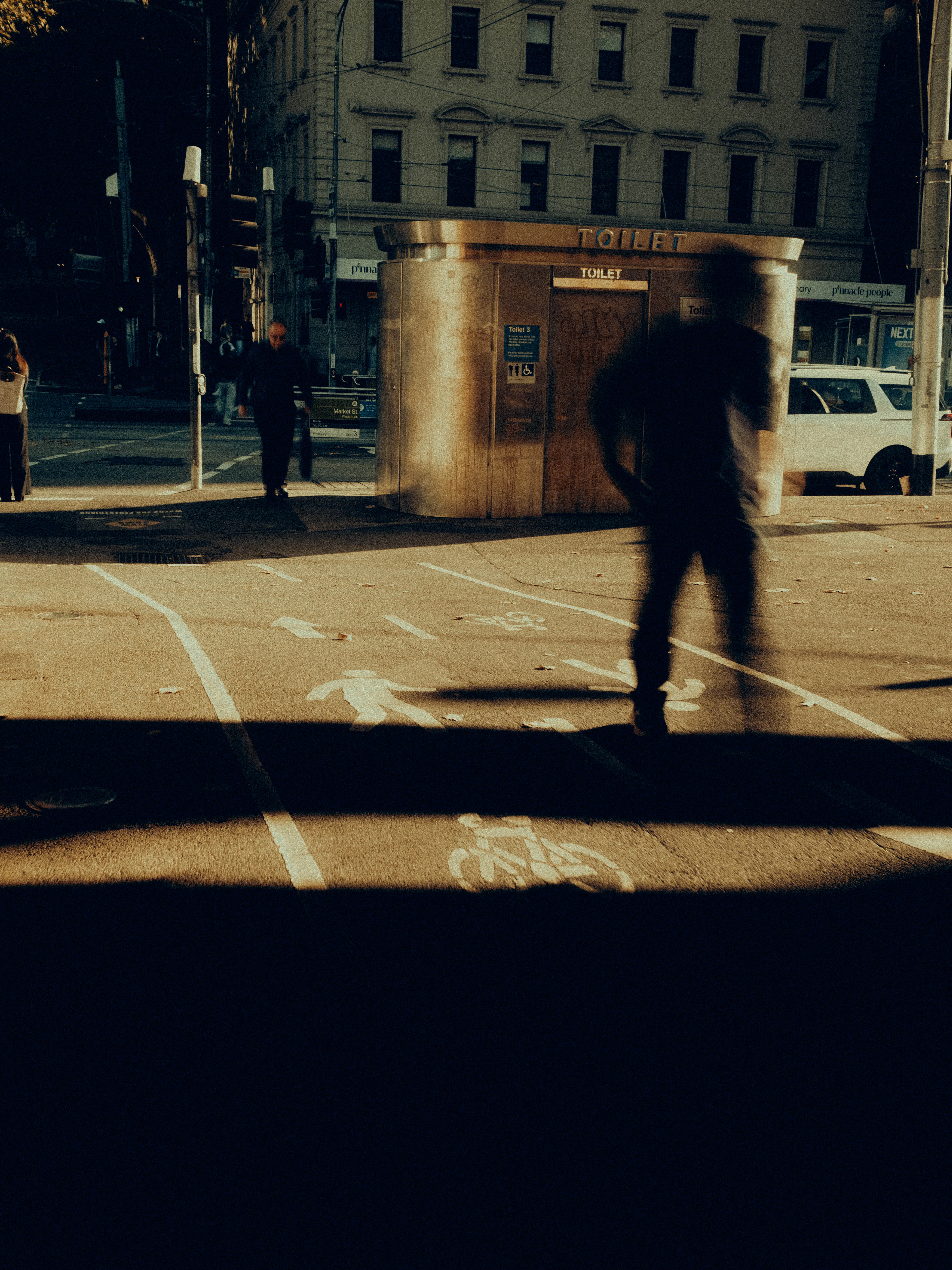 A dark silhouette crosses sunlit Melbourne pavement near a steel public toilet kiosk, long diagonal shadows stretching dramatically across marked bike lanes in the warm midday glare.