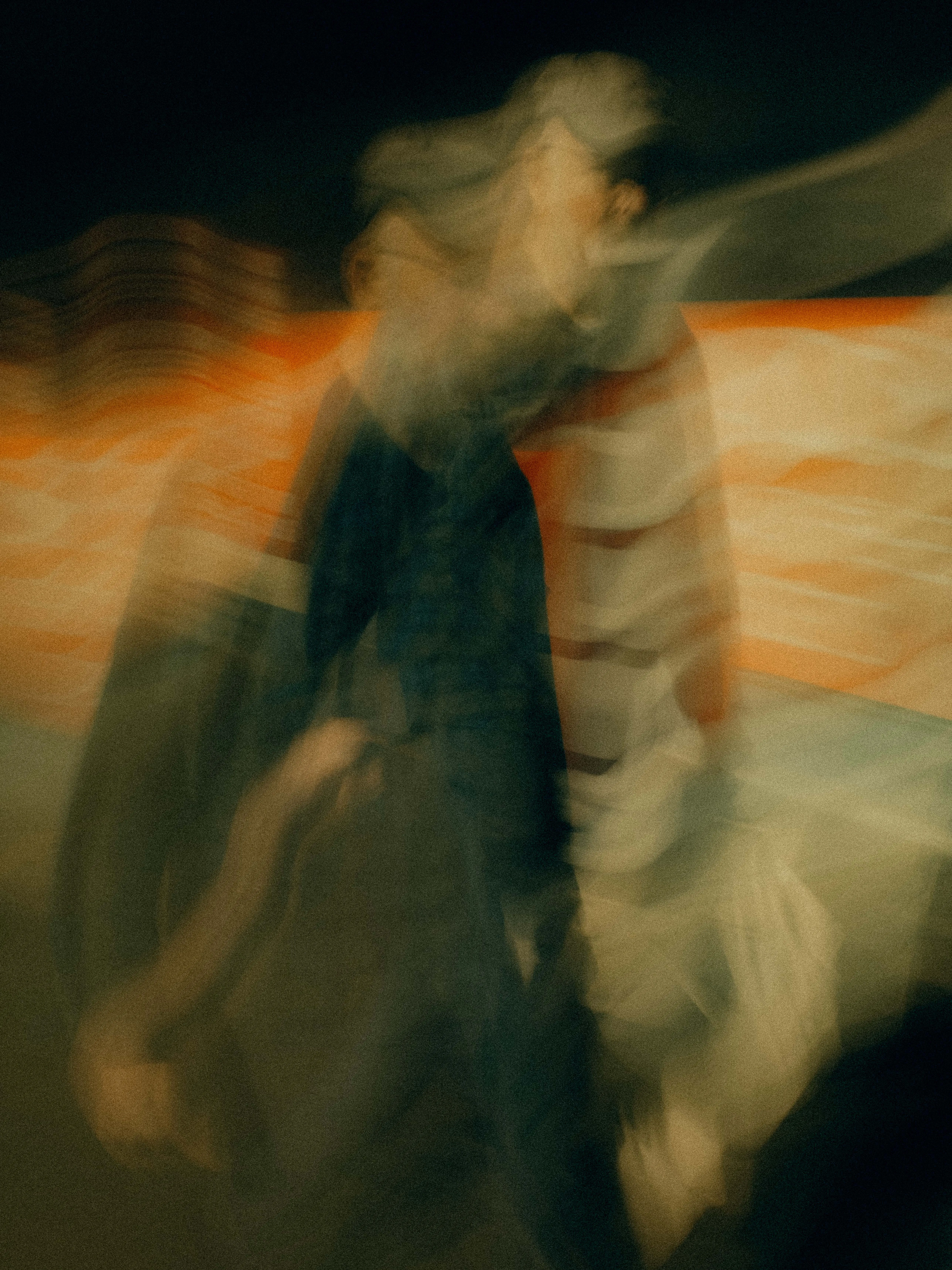 A figure in dark layered clothing drifts through a wash of burnt orange and cream, their body ghosted into overlapping translucent silhouettes against horizontal bands of glowing city light.
