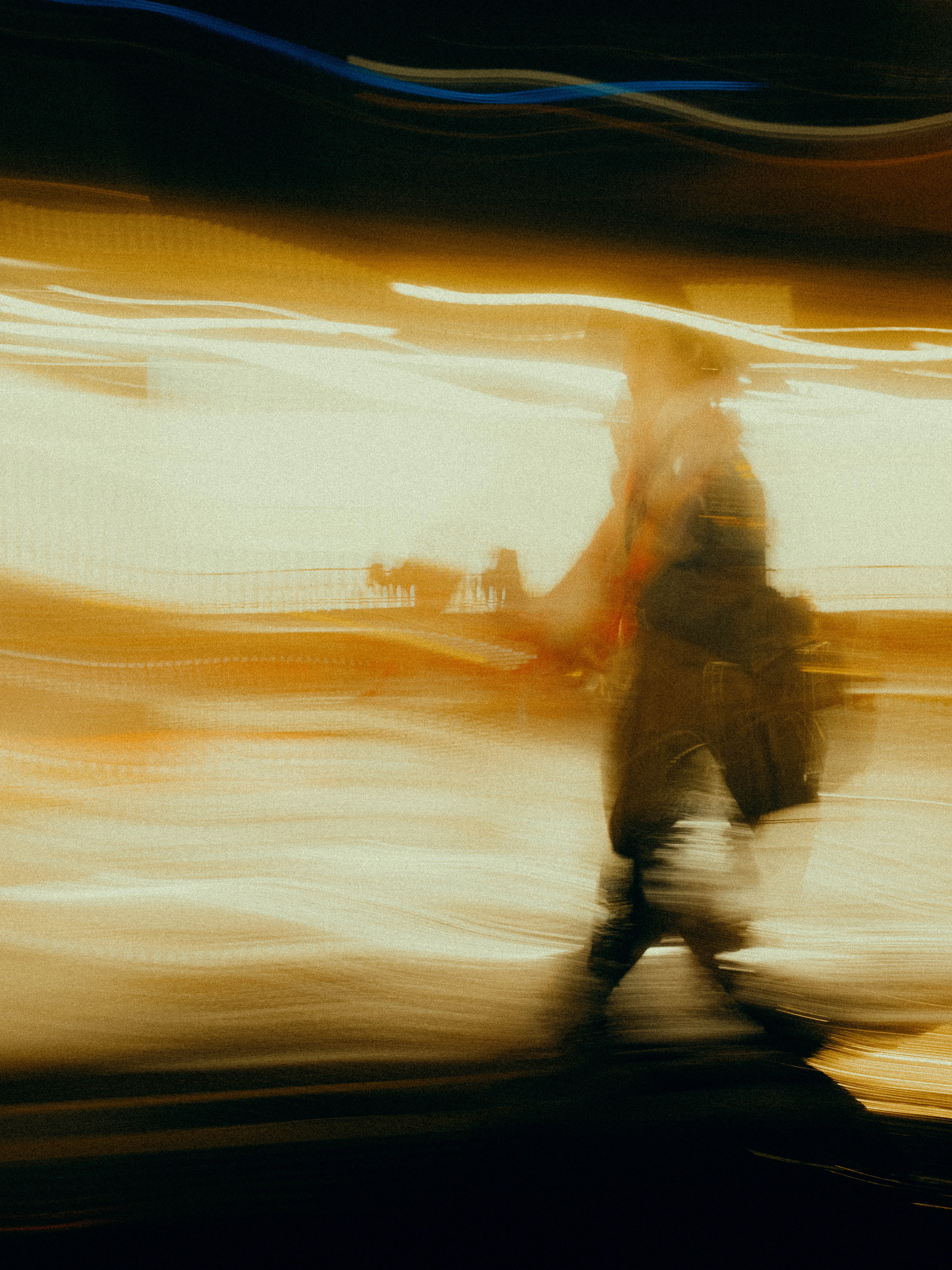 A traveller with luggage crosses a blazing amber transit space, the scene reduced to molten gold and dark silhouette by extreme motion blur and overpowering warm light.